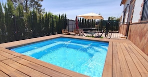 Ein rechteckiger Swimmingpool mit umlaufender Holzterrasse, eine Terrasse mit Stühlen und Sonnenschirm sowie ein Zaun mit Bäumen im Hintergrund.