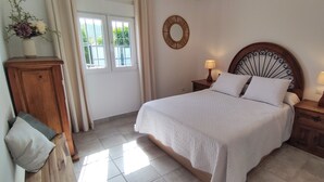 A bedroom with a bed, two bedside tables with lamps, a wooden nightstand, a mirror, and a window with curtains.