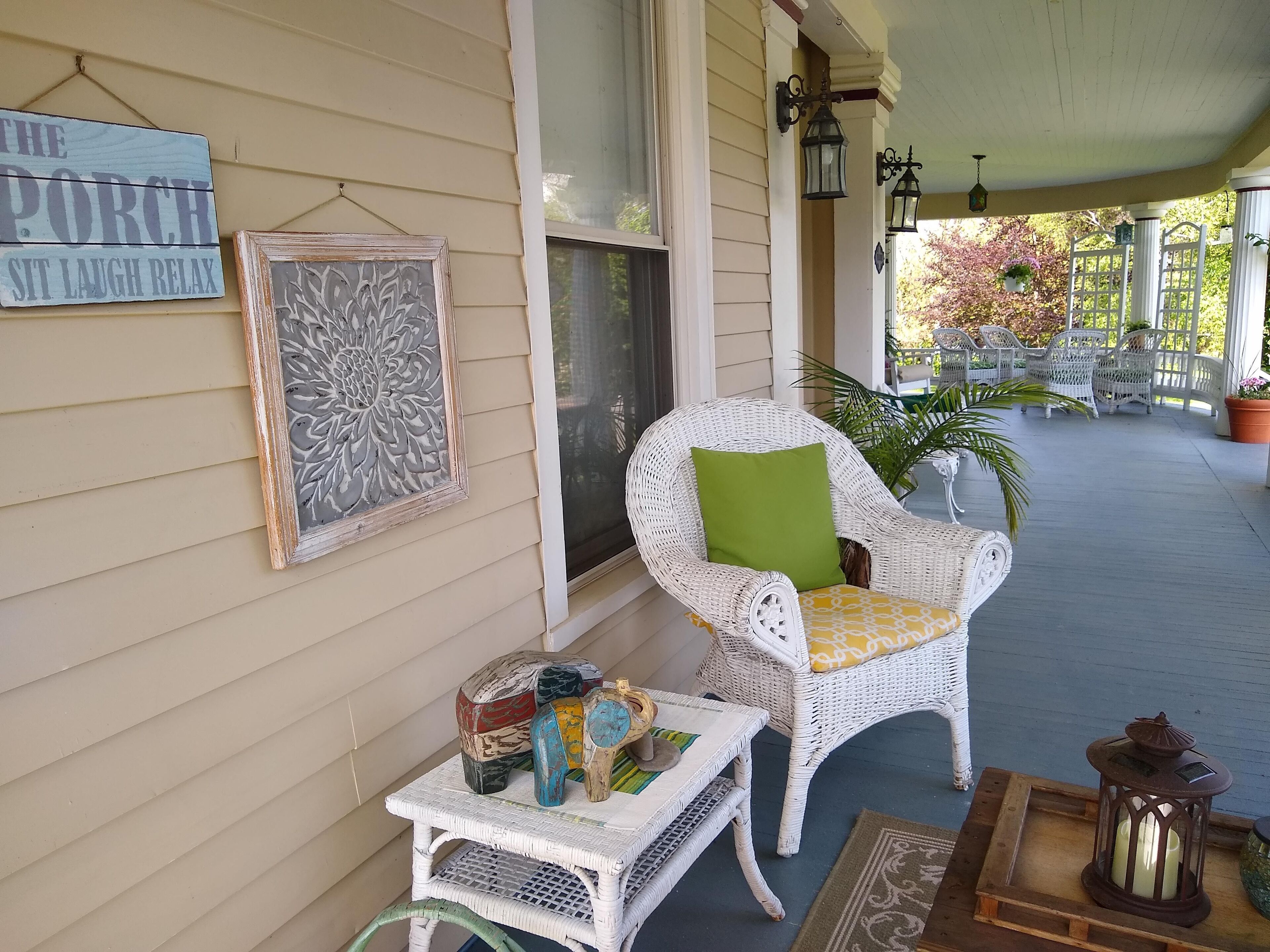 Een veranda met rieten meubels, een klein tafeltje met decoratieve objecten en een bord waarop staat: The Porch Sit Laugh Relax.
