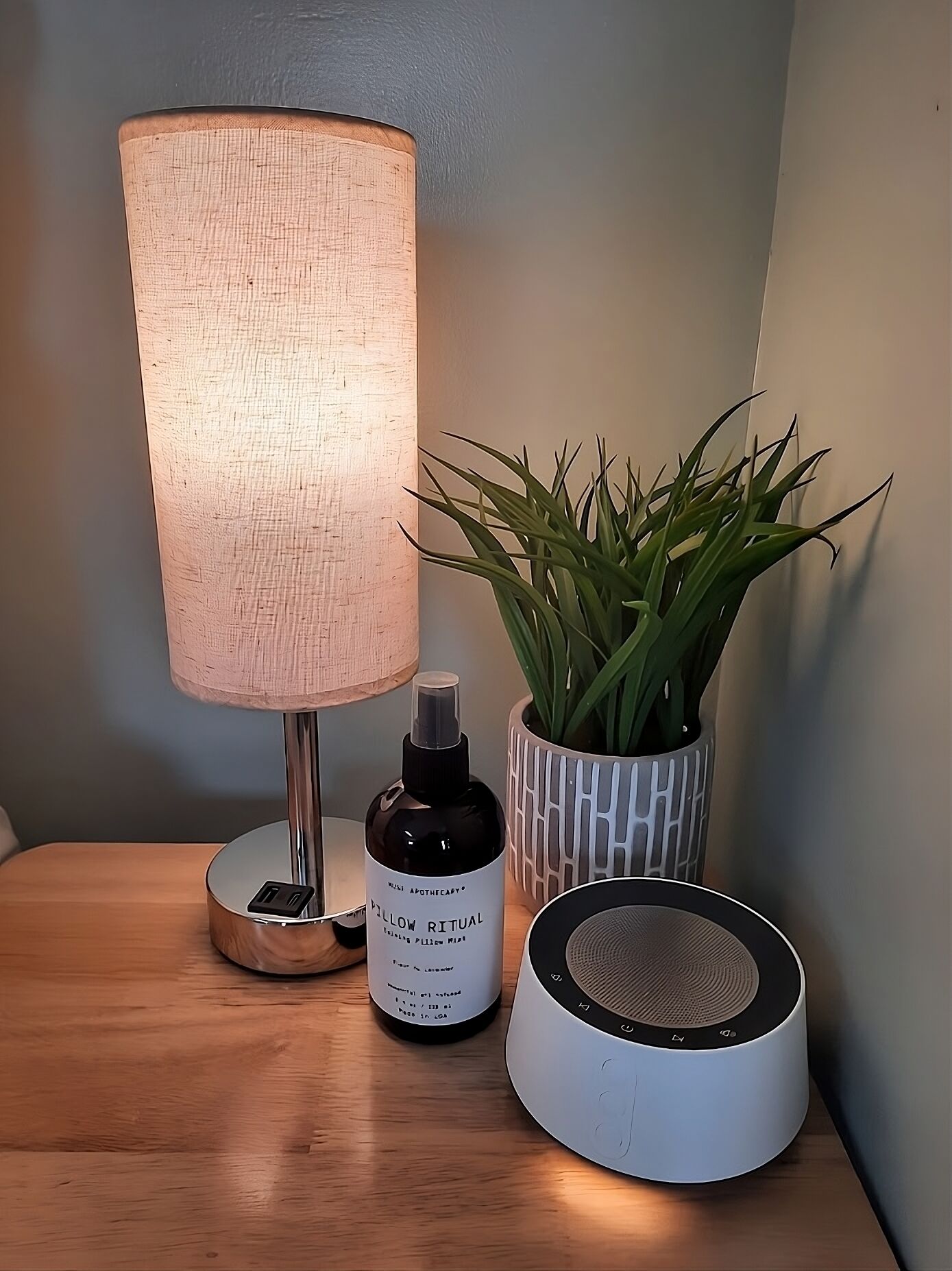 A bedside table with a lamp, a bottle of essential oil, a potted plant, and a speaker.