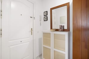 Uma porta branca, um espelho fixado na parede, um telefone e um console de vime com um vaso.