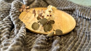 A bed with a grey textured blanket, a wooden tray with dried leaves, and a blue pillow in the background. - Themenhotel Rehlein (Thale)