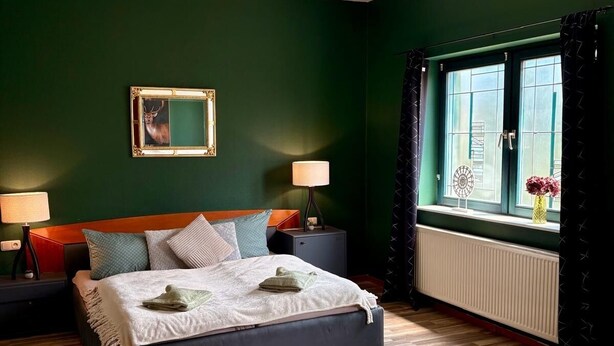 A bedroom with a bed, bedside tables, a window with curtains, a radiator, and a small table with flowers. - Themenhotel Rehlein (Thale)