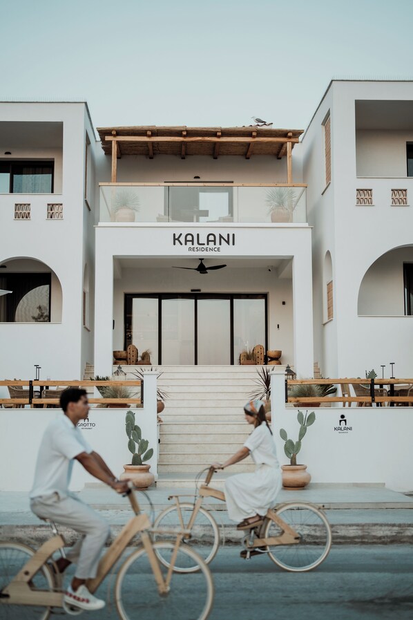 Un bâtiment moderne avec une enseigne indiquant « KALANI RESIDENCE ». On peut voir deux personnes à vélo devant l’entrée.