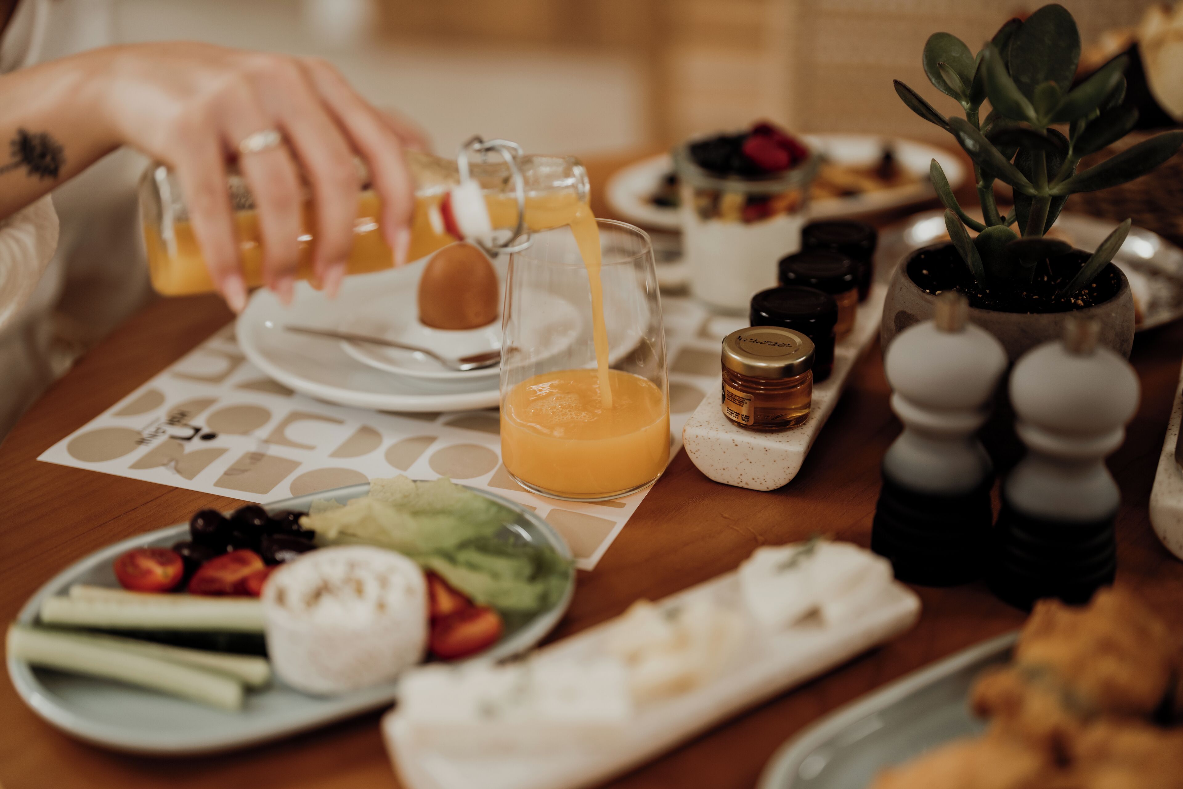 Een ontbijttafel met diverse soorten eten en drinken, waaronder een glas sinaasappelsap, een bord fruit en een kleine potplant.