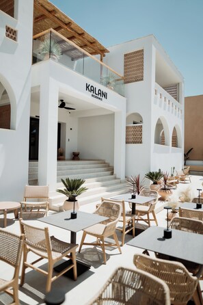 Un bâtiment moderne dont la façade porte l’inscription « KALANI RESIDENCE ». Il dispose d’un espace extérieur aménagé avec des meubles en osier et des plantes en pot.