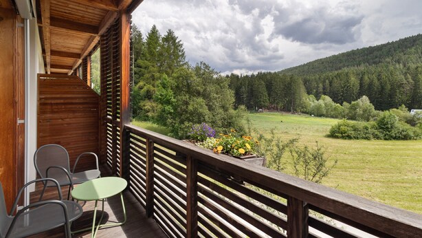 Ein Holzbalkon mit Tisch und Stühlen, der auf eine grüne Landschaft mit Bäumen und einem Hügel blickt.