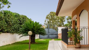 Un jardin bien entretenu, avec un chemin menant à une entrée fermée par un portail, entouré d’une végétation luxuriante et agrémenté de plantes en pot.