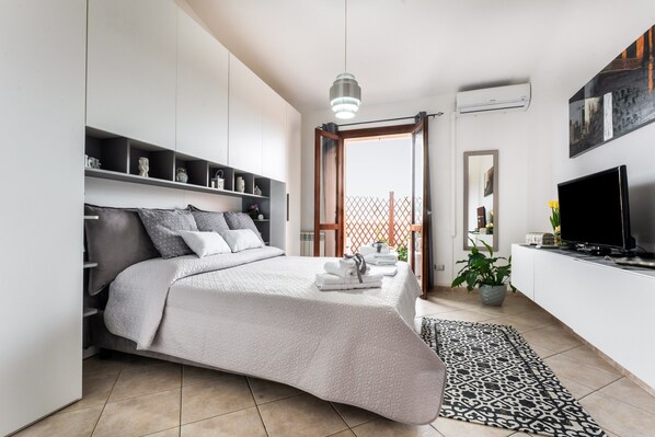 Une chambre moderne avec un lit, une télévision, une plante en pot et une vue sur l’extérieur, visible à travers une porte.