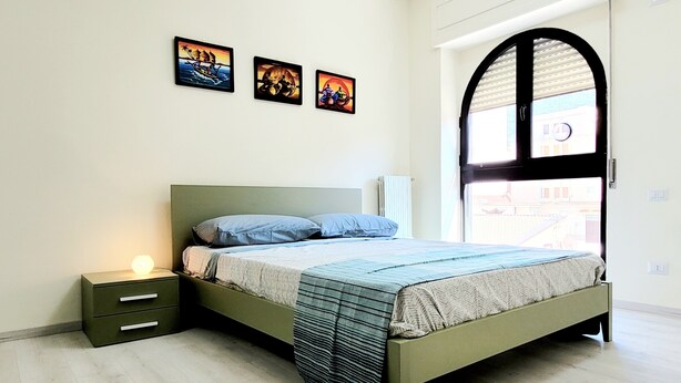 Une chambre à coucher comprenant un lit, une table de chevet, une fenêtre avec des stores et trois tableaux accrochés au mur.