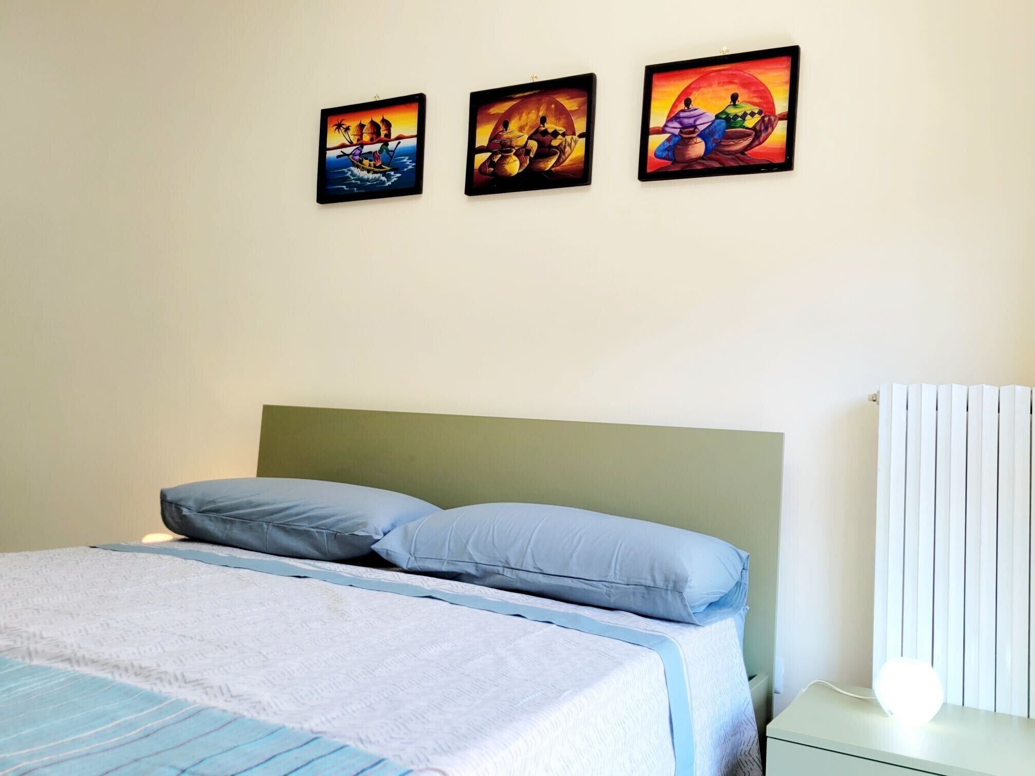 Een slaapkamer met een bed, kussens, een nachtkastje met een lamp en drie aan de muur hangende schilderijen.