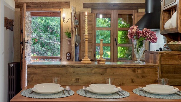 Mesa de madera dispuesta con platos, cubiertos y velas, con vista a follaje a través de una ventana.