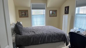 A bedroom with a bed, a desk, two windows with blinds, and two framed pictures on the wall.