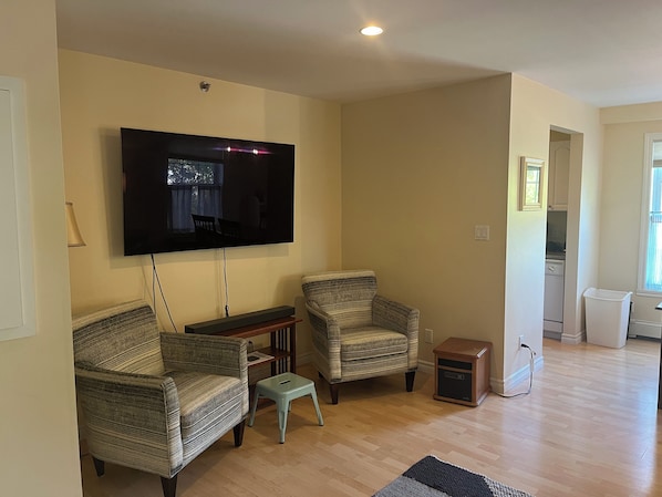 Una sala de estar con un televisor de pantalla plana montado en la pared, dos sillones estampados, un taburete pequeño y un aparador de madera.