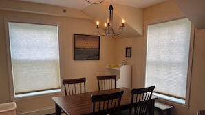 A dining area with a wooden table and chairs, large windows with blinds, a chandelier, and a painting on the wall.