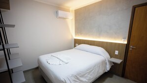 Habitación de hotel ordenada con cama, mesita de noche, aire acondicionado de pared y puerta de madera.
