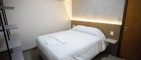 A neatly arranged hotel room with a bed, a bedside table, a wall-mounted air conditioner, and a wooden door.