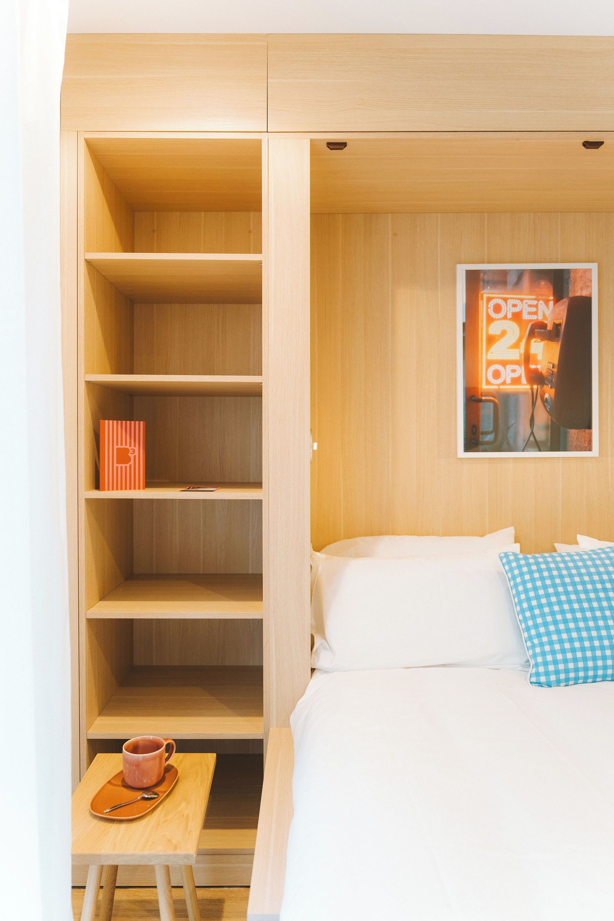 Habitación de hotel con cama, una mesita con taza y platillo, almohada de cuadros y un estante de madera con un cuadro enmarcado.