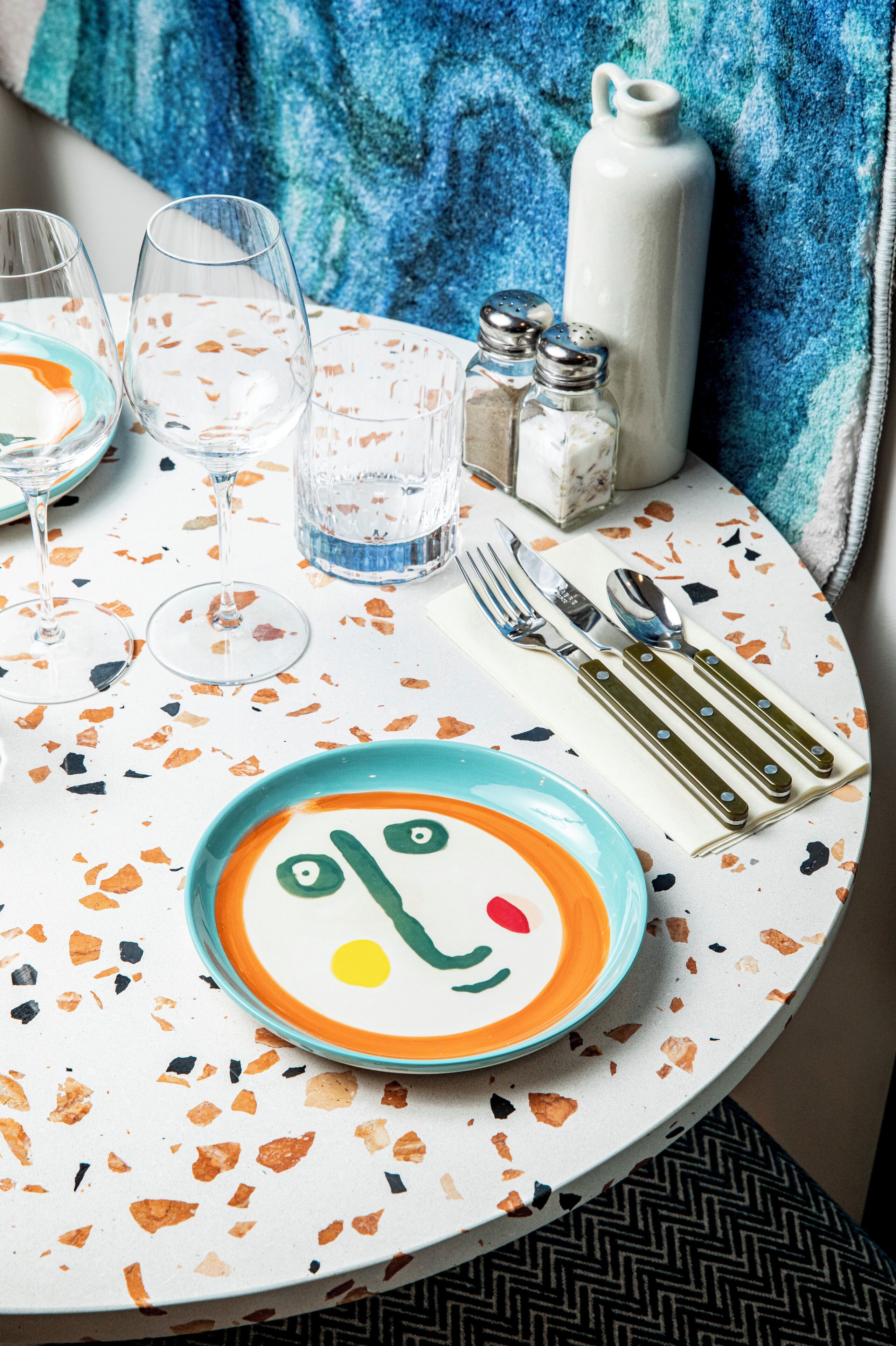 Una mesa redonda con un mantel colorido, un plato con diseño de rostro y un juego de salero y pimentero.