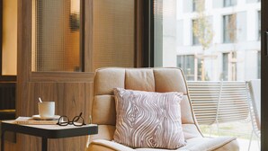 Ein moderner Lounge-Bereich mit gepolstertem Sessel, einem kleinen Tisch mit Tasse und Gläsern sowie einem großen Fenster mit Blick auf Gebäude.