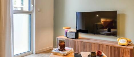 Een moderne woonkamer met een groot raam, een flatscreen televisie, een houten kast en twee kleine tafels met spullen erop.