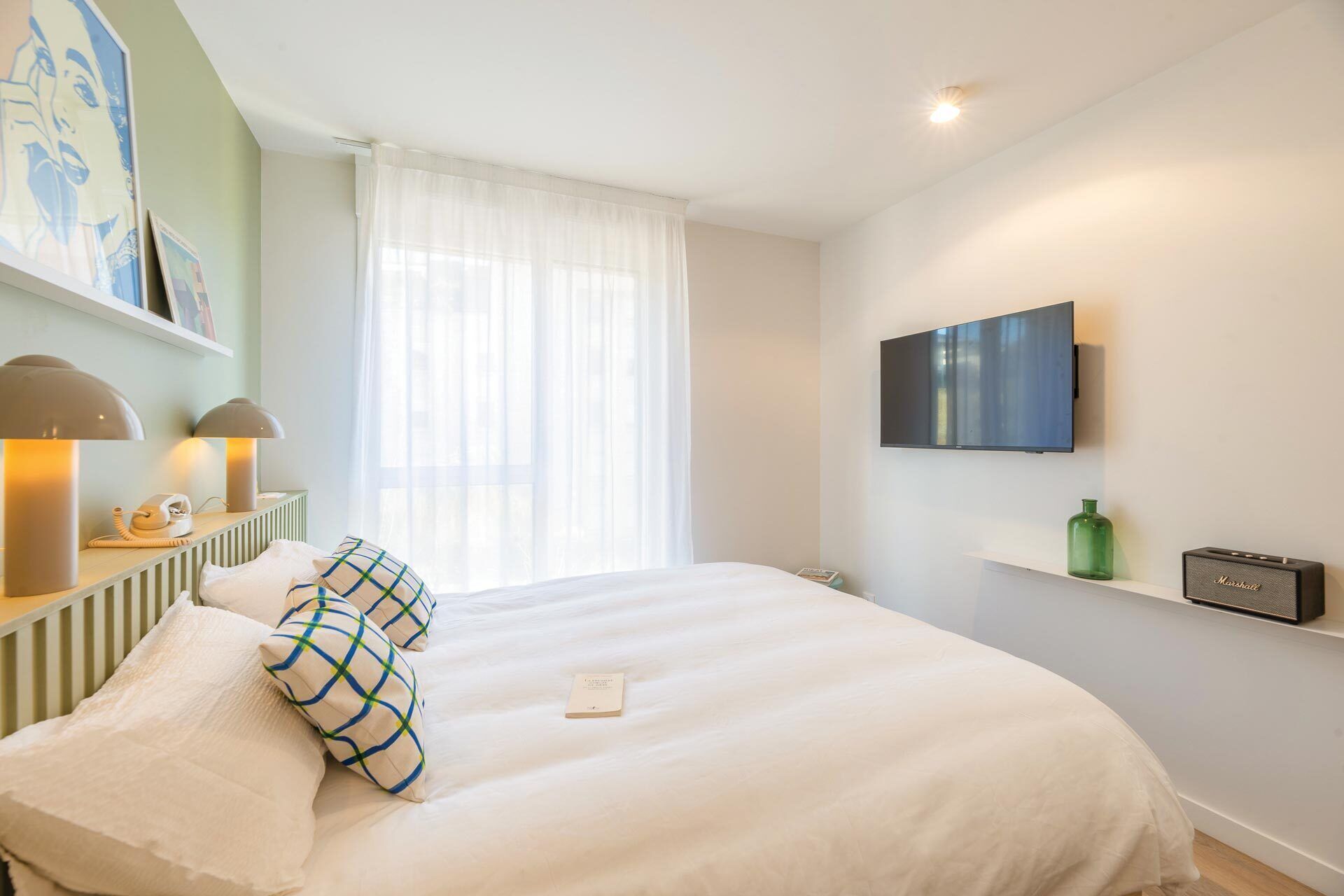 A hotel room with a bed, a television mounted on the wall, a telephone, and a lamp on a bedside table.