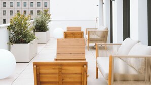 Eine Dachterrasse mit Holzmöbeln, weißen Pflanzgefäßen mit Grünpflanzen und einem modernen Gebäude im Hintergrund.