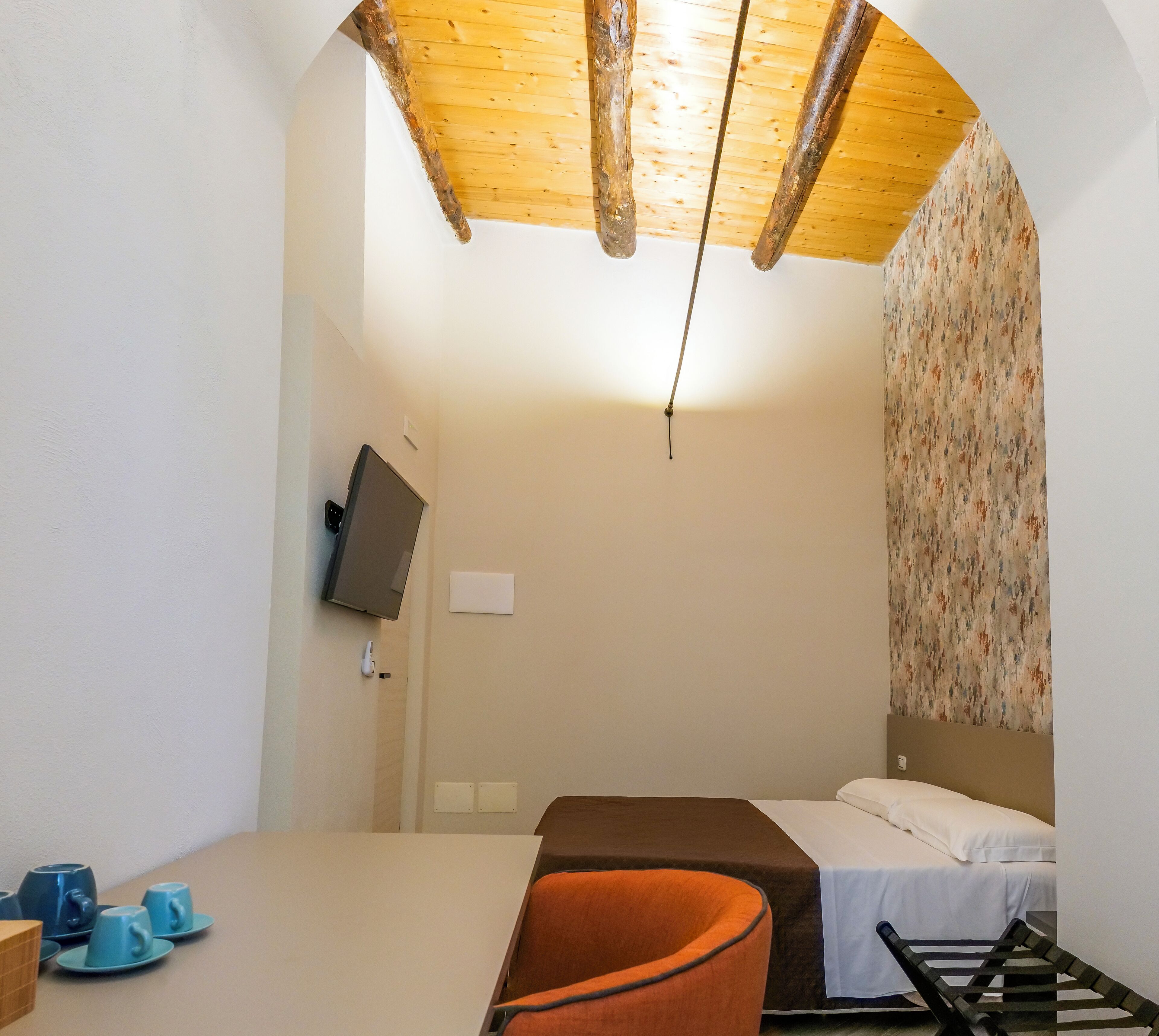A small, cozy room with a bed, a chair, a TV mounted on the wall, and a wooden ceiling.