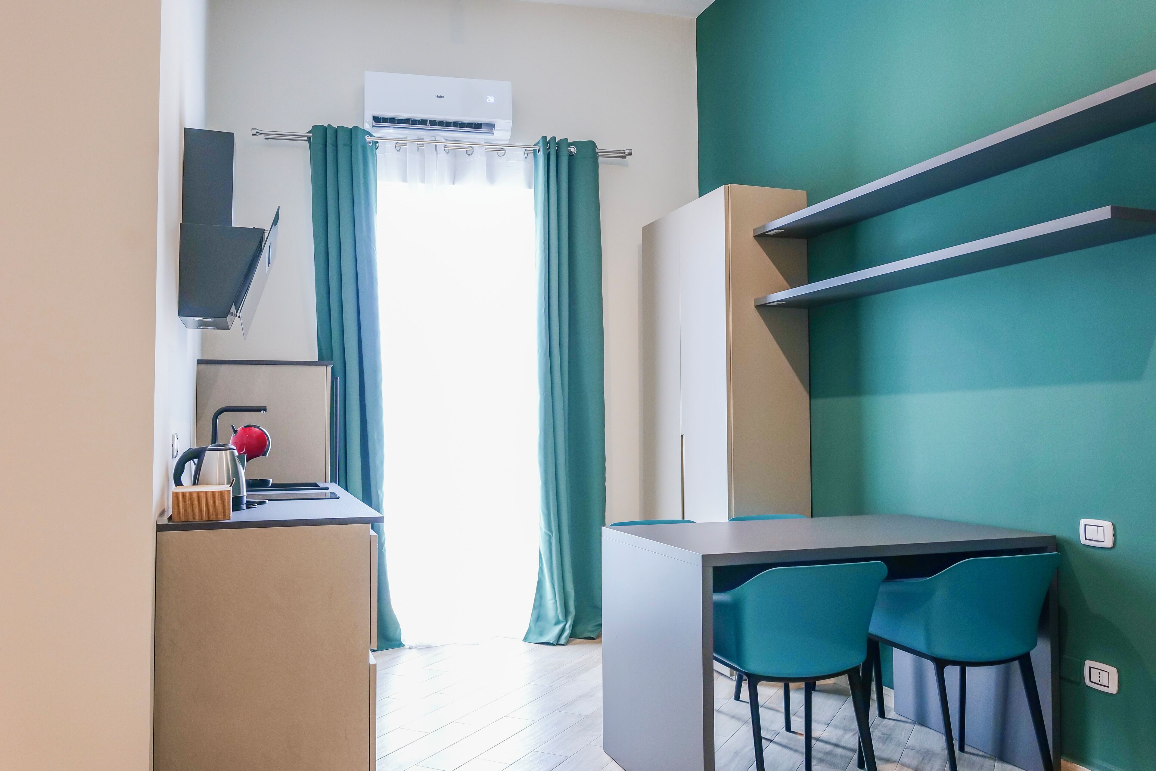 Cocina compacta con lavamanos, hervidor y armarios, un pequeño comedor con dos sillas, y una pared color turquesa.