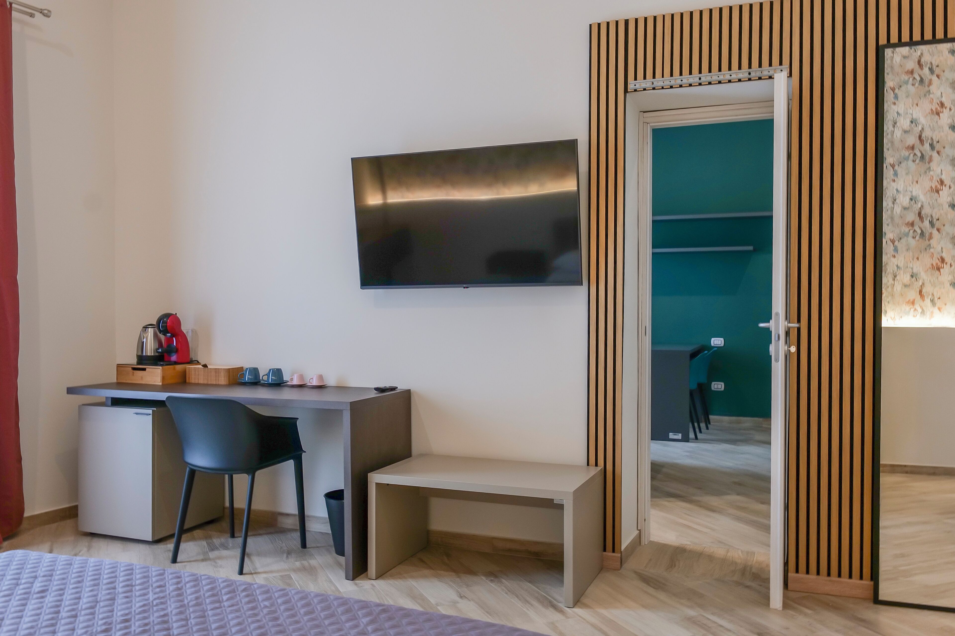 A modern room with a flat-screen TV mounted on the wall, a small desk with a chair, a compact fridge, and a bench.