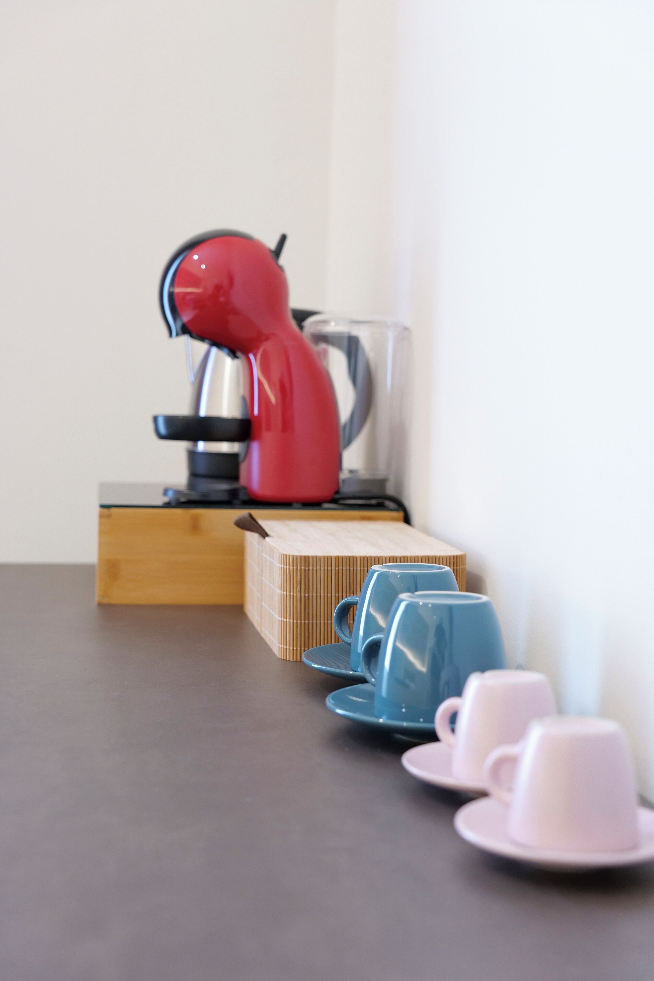 A coffee maker on a wooden box, with two blue cups and two pink cups on a table.