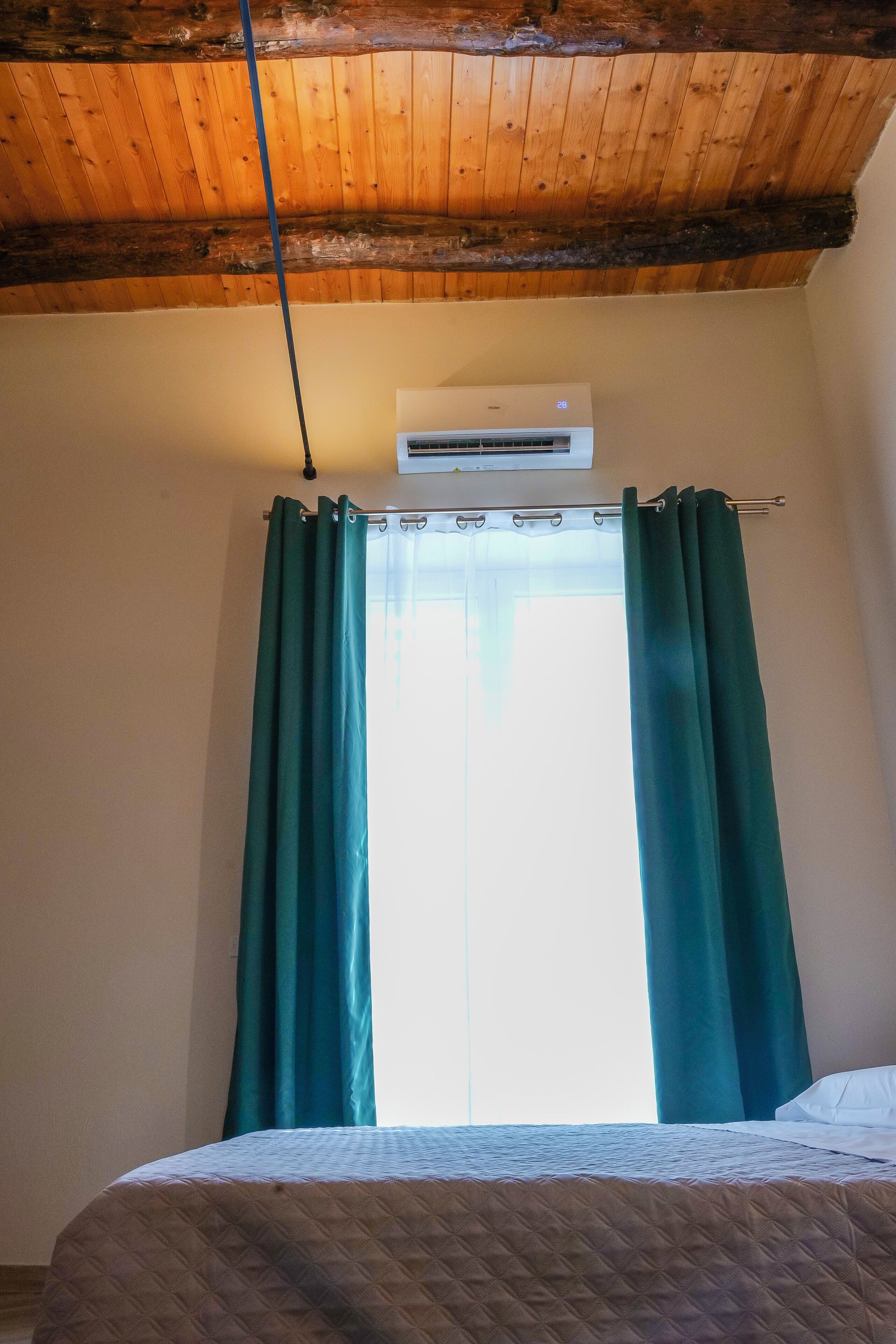 A bedroom with a wooden ceiling, an air conditioning unit, and a window with green curtains.