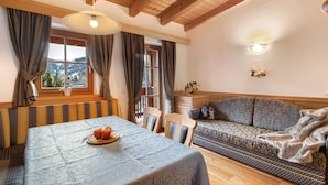 A cozy room with a wooden ceiling, a dining table with a patterned tablecloth, a sofa with patterned cushions, and a window with curtains.