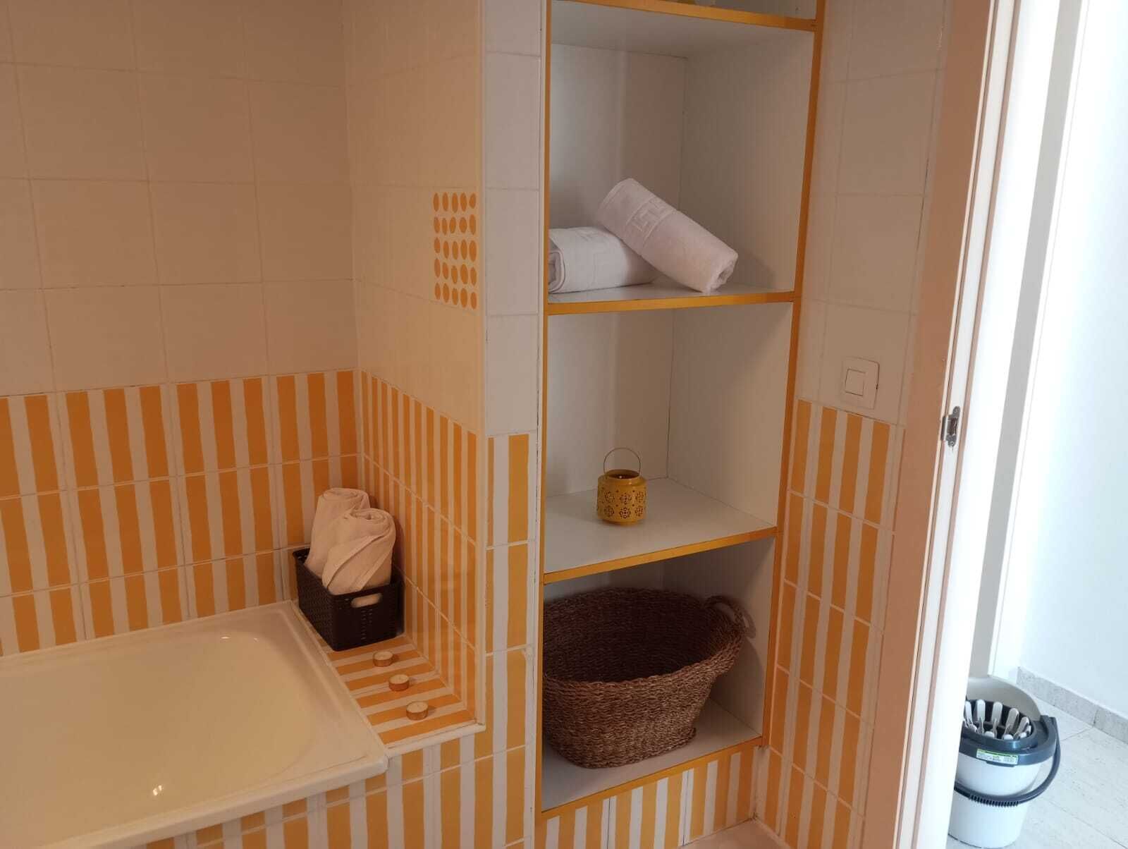 Une salle de bain avec des murs à rayures, une baignoire blanche, une étagère avec des serviettes et un panier, ainsi qu'une poubelle.