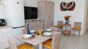 A modern kitchen with a dining area, featuring a large flat-screen TV, a white refrigerator, wicker chairs with yellow cushions, and a decorative wall piece.