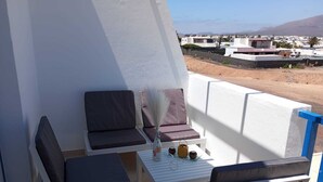 A balcony with two chairs and a small table, a vase with flowers, and a view of a coastal town.