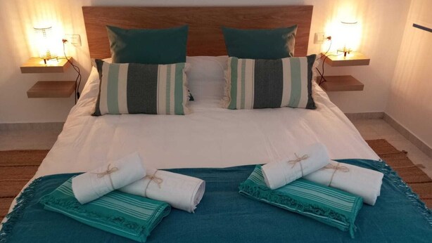 A neatly made bed with a wooden headboard, two bedside tables with wall-mounted lamps, and two rolled towels on the bed.