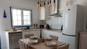 A modern kitchen with white cabinets, a stainless steel dishwasher, a white refrigerator, and a dining area with wicker chairs and a wooden table.