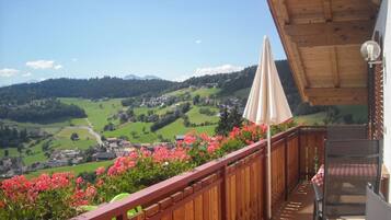 Un balcone con ringhiera in legno, un tavolo con tovaglia a scacchi e vista su una valle verde con case e montagne sullo sfondo.