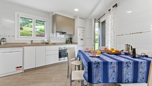A modern kitchen with white cabinetry, a dining table with a blue tablecloth, and a view of greenery outside. - Country House 'Casa La Manzana' with Mountain View, Private Terrace and Wi-Fi (Villaviciosa)