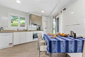 A modern kitchen with white cabinetry, a dining table with a blue tablecloth, and a view of greenery outside.