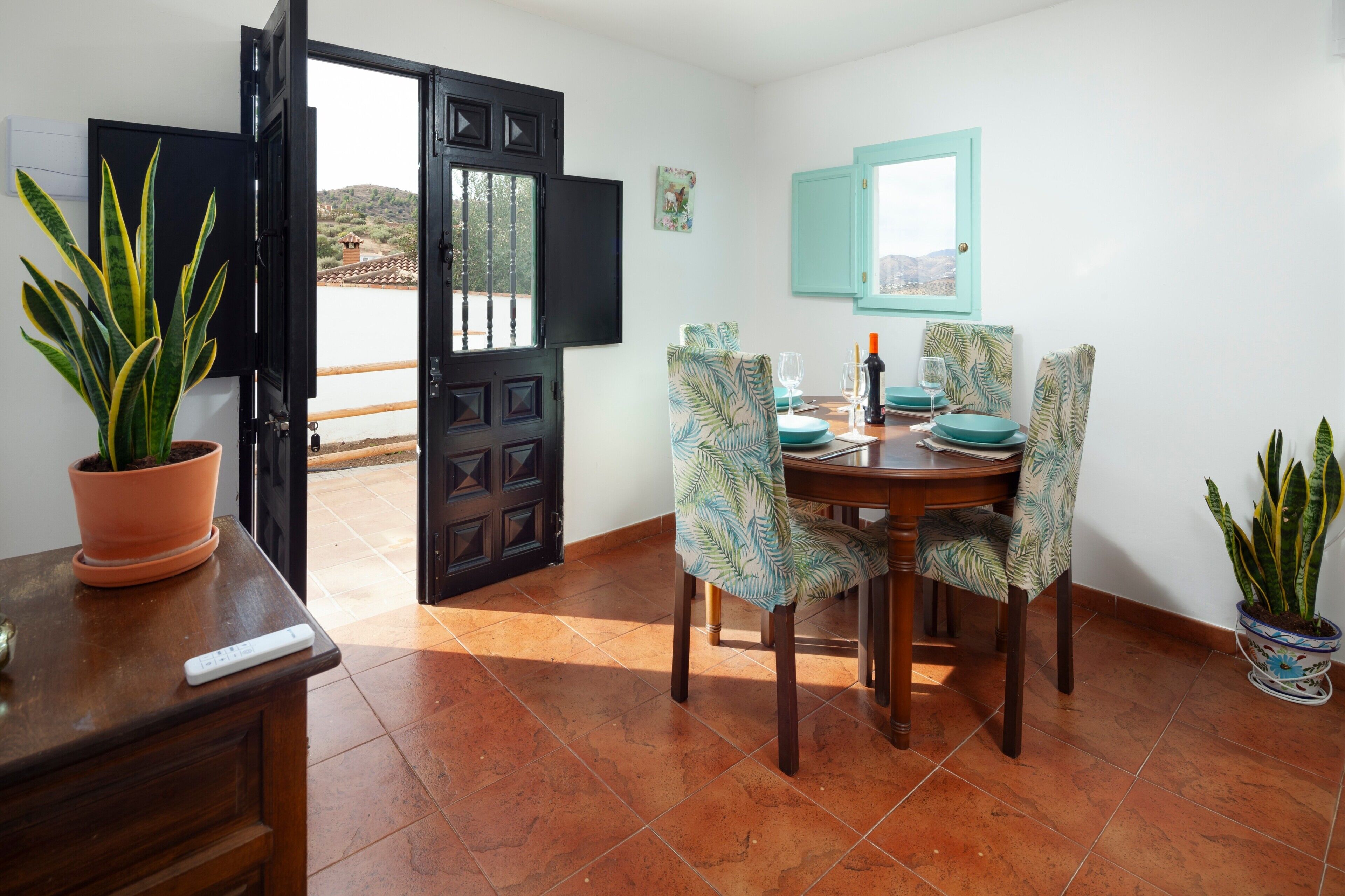 Comedor con una mesa dispuesta para dos, sillas con estampado de hojas tropicales, una planta en maceta sobre la mesa y vistas a través de un ventanal abierto.