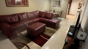 Uma sala de estar com um sofá seccional de couro marrom, uma mesa de jantar com cadeiras, um ventilador de teto e um quadro na parede.