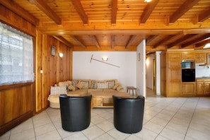 A wooden interior with a sofa, two black stools, a wall clock, and wooden cabinets.