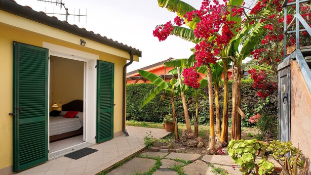 A yellow building with green shutters, a bed, and a small garden with red flowers and plants. - Villa 'Pairola' with Sea View, Private Garden and Air Conditioning (San Bartolomeo al Mare)