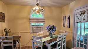 A dining area with a wooden table, white chairs, a vase with flowers, a stained glass window, and a door with a floral curtain. - YEAR'ROUND FAMILY FUN! (Warrensburg)