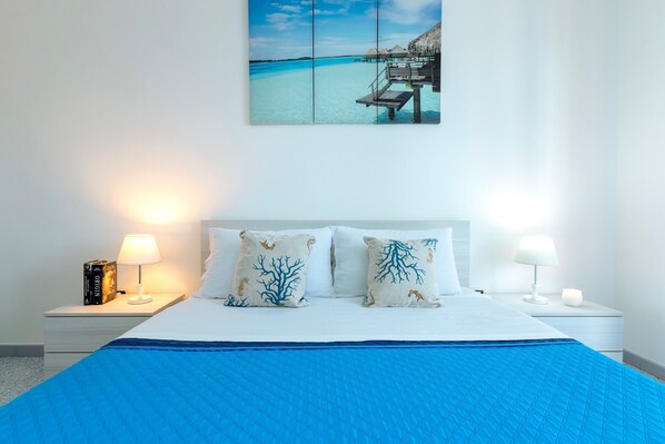 A bed with a blue quilt, white pillows with coral tree designs, a nightstand with a lamp, and a wall-mounted picture of a beach.