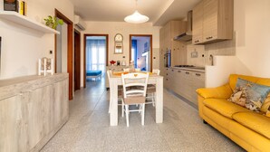 A modern kitchen with wooden cabinets, a dining area with a white table and chairs, and a yellow sofa with a decorative pillow. - Apartment 'Terrace with Sea View and Barbecue' with Sea View, Wi-Fi and Air Conditioning (Alghero)