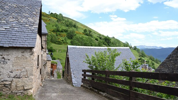 Un étroit sentier de pierre mène à des bâtiments traditionnels en pierre, coiffés de toits d’ardoise, et est entouré de collines verdoyantes et d’un ciel dégagé.