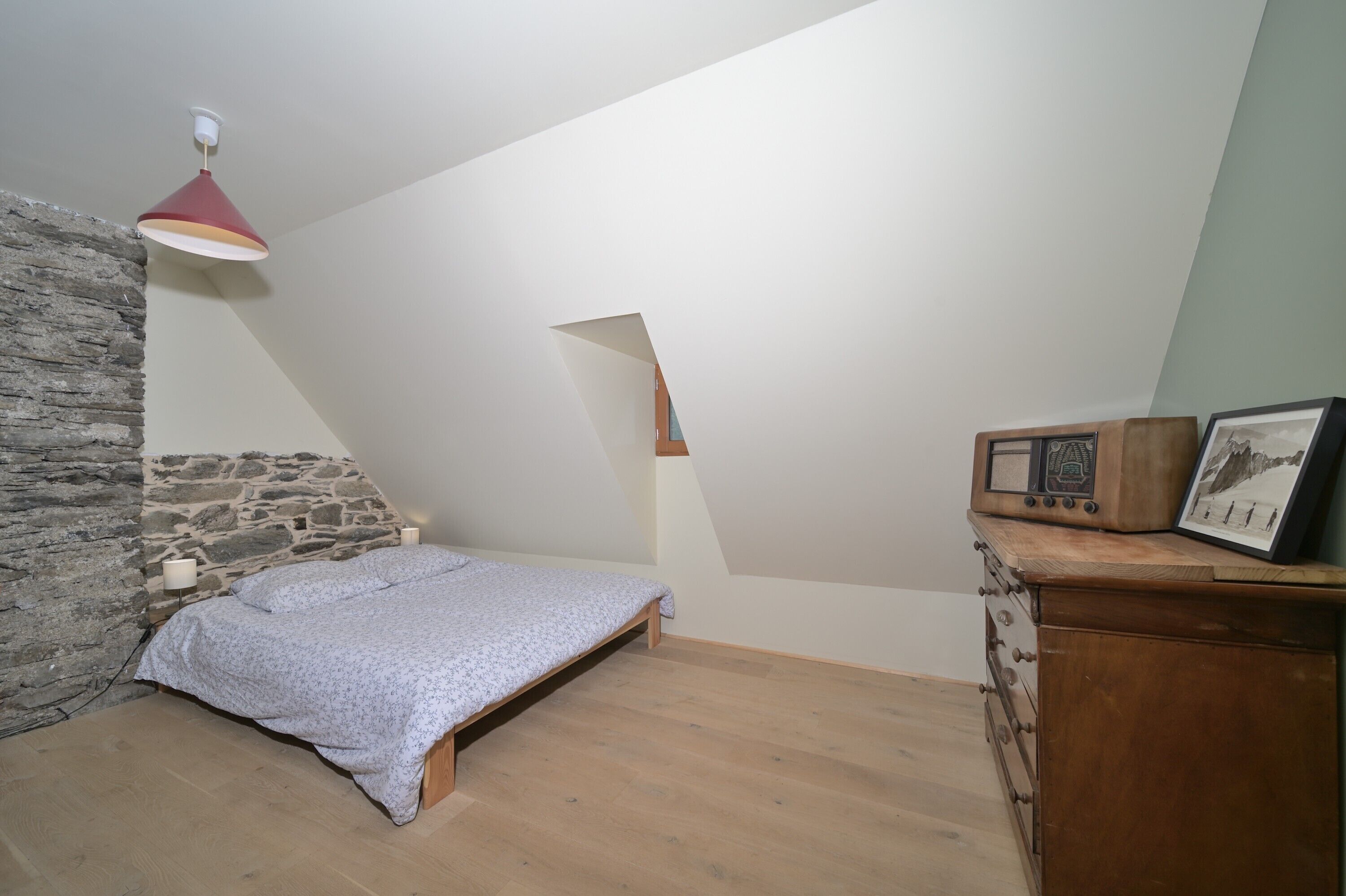 A bedroom with a sloped ceiling, a bed, a dresser, and a stone wall.